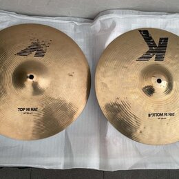 Dva mosazné činely typu hi-hat položené na bílé podložce. Horní činel má nápis TOP HI HAT a logo Zildjian. Spodní činel má nápis BOTTOM HI HAT a výra…