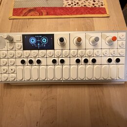 Bílý kompaktní synthesizer s klávesou a ovládacími prvky. Na pravém boku je svisle napsáno OP-1 field. Zařízení obsahuje displej, čtyři barevné otočn…