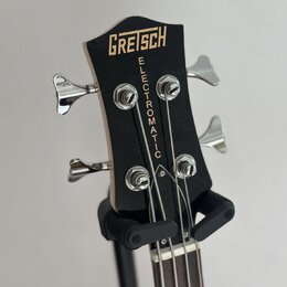 Černá hlava basové kytary s nápisem GRETSCH v horní části a svislým nápisem ELECTROMATIC pod ním. Nástroj má čtyři chromované ladičky. Povrch je hlad…