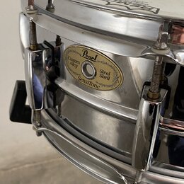 Detailní pohled na kovový snare drum stříbrné barvy s chromovaným obručováním. Na korpusu je oválný zlatý štítek s nápisy Pearl, Custom Alloy, Steel…
