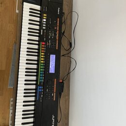 Černý digitální syntezátor s klaviaturou, umístěný na dřevěném stole. Na přístroji jsou viditelná loga a nápisy Roland a JUPITER-80. Ovládací panel o…