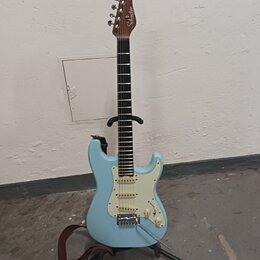 Světle modrá elektrická kytara s bílým pickguardem a hnědým hmatníkem. Na hlavě je bílý nápis "Squier". Nástroj stojí na černém stojanu a je k němu p…