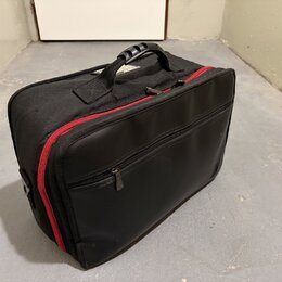 Černý transportní kufr z textilního a syntetického materiálu s červeným zipem. Má dvě úchytky, jednu plastovou a jednu textilní. Na přední straně je…