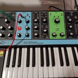 Semi-modulární analogový syntetizátor s klaviaturou a ovládacím panelem v různých barvách (šedá, modrá, zelená). Na spodní liště je nápis GRANDMOTHER…