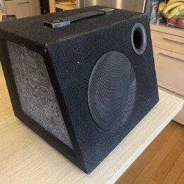 Černý kostkový subwoofer potažený texturovaným materiálem s madlem na horní straně. Přední strana obsahuje kruhový reproduktor krytý černou síťkou a…