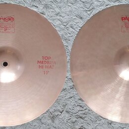 Dva mosazné činely typu hi-hat položené vedle sebe na bílém koberci. Oba mají červené nápisy a logo Paiste 2002. Levý činel má nápis TOP MEDIUM HI-HA…