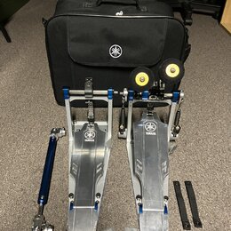 Sada kovových bubnových pedálů v modré a stříbrné barvě s logem YAMAHA. Součástí je černá přepravní taška s bílým logem Yamaha, dvě žluté gumové podl…