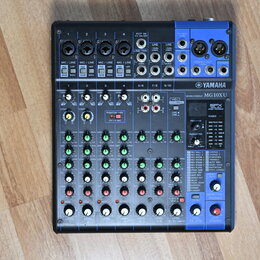 Černý mixovací pult s modrými prvky a bílými nápisy. Na přístroji je viditelné logo YAMAHA a označení modelu MG10XU MIXING CONSOLE. V pravé části se…