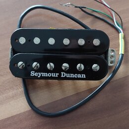 Černý humbucker s kovovými póly a bílým nápisem Seymour Duncan. Nástroj má připojený kabel s několika barevnými vodiči. Povrch je lesklý a vykazuje m…