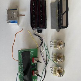 Sada komponentů pro elektrickou kytaru rozložená na bílém povrchu. Obsahuje černý ovladač s nápisem SUSTAINIAC®, černý blok s dvěma červenými snímači…