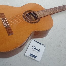 Kytara s přírodním dřevěným povrchem a hnědým hmatníkem. Vedle nástroje leží krabička s nápisy Blond STRINGS, classical guitar Medium a nečitelný tex…