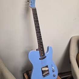 Modrá elektrická kytara s tmavým hmatníkem, umístěná na stojanu. Na hlavě je viditelné logo Fender. Nástroj má jeden viditelný snímač u mostu, jeden…