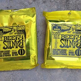 Dvě žluté fóliové balení kytarových strun. Na obou je nápis ERNIE BALL BEEFY SLINKY, CUSTOM GAUGE 3627, NICKEL WOUND MADE GUITAR STRINGS IN USA a ozn…