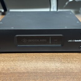 Černý kovový přístroj s hranatým tvarem. Na přední straně je nápis UAD-2 Satellite a displej s logem UA a textem UNIVERSAL AUDIO a HOST. Povrch je ma…