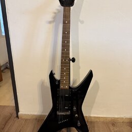Černá elektrická kytara s ostře hranatým korpusem umístěná na stojanu. Na hlavě hmatníku je bílé logo Jackson. Nástroj má dva humbuckery, z nichž na…