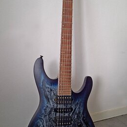 Elektrická kytara s modrým průsvitným povrchem a viditnou strukturou dřeva. Na hlavě hmatníku je bílý nápis Ibanez. Nástroj má dva humbuckery, jeden…