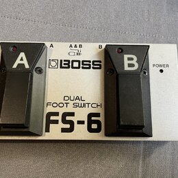 Stříbrný dvoupedálový spínací jednotka s černými plasty. Na přístroji jsou nápisy BOSS, DUAL FOOT SWITCH a FS-6 oraz označení pedálů A a B. V dolní č…
