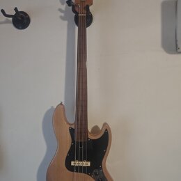 Elektrická basová kytara s přírodním dřevěným povrchem těla a hředu. Nástroj má černý pickguard, dva snímače a čtyři ladicí kolíky na hlavě. Na těle…