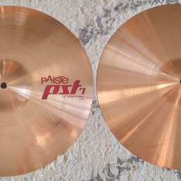 Dvě mosazné činely typu hi-hat položené vedle sebe na šedém koberci. Levý činel nese nápisy pAiste, pst 7 a 14" HEAVY HI-HAT TOP. Pravý činel má nápi…