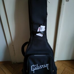 Černý softcase na kytaru s bílým nápisem Gibson v dolní části. Na popruhové části je připevněna bílá papírová štítková visačka s nečitelným textem. P…