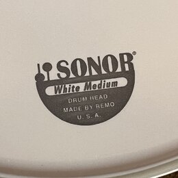 Detail bílého bubnového koženě s černým potiskem. Je na něm vidět logo SONOR a nápisy White Medium, DRUM HEAD, MADE BY REMO a U. S. A. Povrch je hlad…