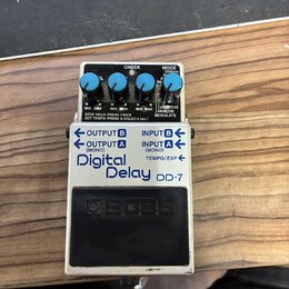 Stompbox efekt s bílým kovovým šasi a černým spínacím pedálem. Na přední straně je nápis Digital Delay DD-7 a logo BOSS. Zařízení má čtyři modré ovlá…