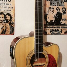Akustická kytara s přístupem pro praváky, přírodní barva těla a tmavě hnědá hlava. Na hlavě je viditelné logo Epiphone. Kytara má červený pickguard a…