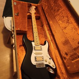 Černá elektrická kytara v hnědém tvrdém kufru s oranžovou výplní. Nástroj má bílý pickguard, tři snímače a světle hnědý javorový hриф. Na hlavě kytar…