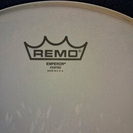 Detail bílého, matného bicího koženu. Uprostřed je černé logo REMO v podobě koruny a pod ním nápisy EMPEROR®, COATED a MADE IN U. S. A. V dolní části…