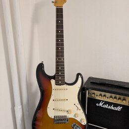 Elektrická kytara s tónem sunburst, bílým pickguardem a javorovou hlavou. Na hlavě je viditelné logo Fender. Vedle kytary stojí černý kytarový zesilo…