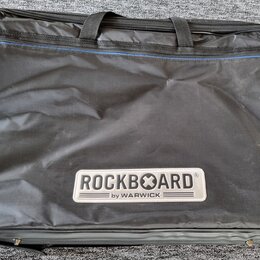 Černý textilní pouzdro na hudební nástroj s dvěma rukojtěmi a zipem. V dolní části je bílý obdélníkový štítek s nápisem ROCKBOARD® a pod ním menší te…