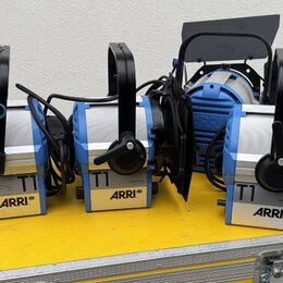 Tři modré studiové reflektory s černými klapkami a stříbrnými prvky, umístěné na žlutém transportním kufru. Na každém z přístrojů je viditelný nápis…