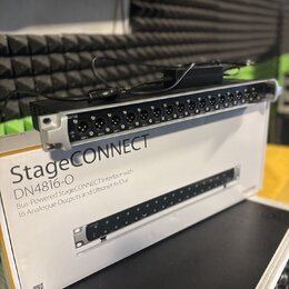 Na fotografii je stříbrno-černý rackový audio rozhraní s řadou 16 XLR konektorů. Zařízení leží na bílé krabici s nápisy StageCONNECT, DN4816-O a text…