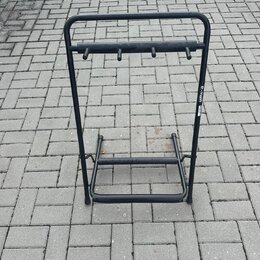 Černý kovový stojan na hudební nástroje s horní lištou vybavenou čtyřmi kolíky. Na pravém bočním sloupeku je viditelný bílý nápis th.mann. Povrch sto…