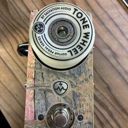 Kytarový efekt s bílým ovládacím knoflíkem a šasi z texturovaného materiálu v hnědo-šedých tónech. Na knoflíku je nápis BY SALVATION AUDIO TONE WHEEL…