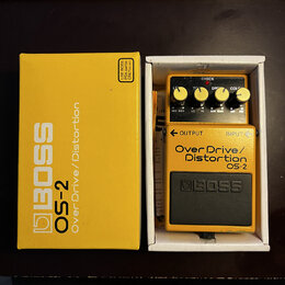 Žlutý kytarový efekt BOSS OS-2 Over Drive/Distortion v originální žluté krabici. Na pedálu jsou čtyři ovládací knofíky a nápisy CHECK, TONE, DRIVE a…