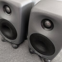 Dva šedé studiové monitory s matným povrchem a zaoblenými hranami. Na spodní části obou reproduktorů je bílý nápis GENELEC. Každý monitor má v horní…