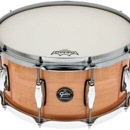 Kytarový snare drum s dřevěným korpusem v přirozené hnědé barvě a chromovaným obručováním. Na korpusu je oválný černý štítek s bílým nápisem Gretsch…