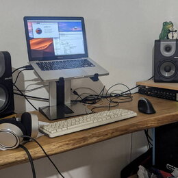 Na dřevěném stole je umístěno studio s notebookem na stojanu, dvěma černými studiovými monitory s nápisem M-Audio a bílou klávesnicí. V popředí leží…