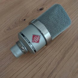Stříbrný kondenzátorový mikrofon s kovovou síťovkou. Na těle je nápis NEUMANN MICROPHONE a červenobílý štítek s nečitelným nápisem. Na spodní části j…