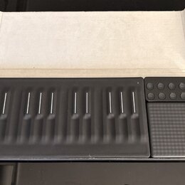 Černý MIDI kontrolér s gumovou klávesnicí a doprovodným touchpad-padem v bílém kartonovém obalu. Zařízení obsahuje řadu vyvýšených kláves s bílými vo…