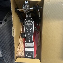 V kartónové krabici se nachází červený bubnový pedál. Na horní části je bílý oválný štítek s nápisem DRUMS STAND a pod ním Stabla. Pedál má kovové me…