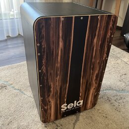 Černý cajon s přední deskou v imitaci tmavého dřeva. Ve spodní části je bílý nápis Sela a pod ním menší text the soul of sound. Povrch je hladký, v r…