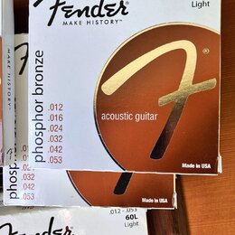 Fotografie zobrazuje tři krabičky strun pro akustickou kytaru. Na obalech je bílé pozadí, hnědý kruh se zlatým logem „F“ a nápisy „Fender“, „MAKE HIS…