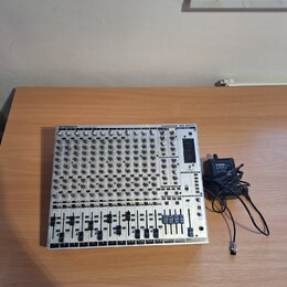 Stříbrný mixážní pult s černými posuvníky a ovládacími prvky. V horní části je uveden nápis BEHRINGER a model EURORACK MX 2642A. Vedle přístroje leží…