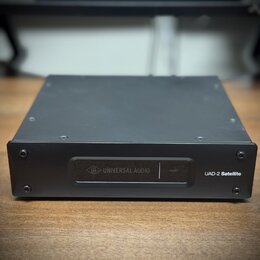 Černý kovový přístroj obdélníkového tvaru. Na přední straně je nápis UAD-2 Satellite a panel s logem a názvem UNIVERSAL AUDIO oraz nápisem HOST. Povr…