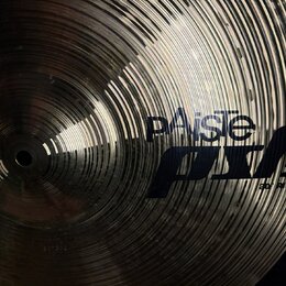 Detailní pohled na kovový činel s koncentrickými rýhami. Na povrchu je černý nápis „paiste“ a vedle něj velké logo „ PST 7“ s doplňkem „RD RIDE“. V d…