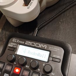Černý kytarový procesor s nápisy ZOOM G1X FOUR a Multi-Effects Processor. Na displeji je zobrazen text 24 RocknRoll. Zařízení je připojeno k bílému n…