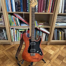 Elektrická kytara s dřevem v přirozené hnědé barvě, černým pickguardem a černými potenciometry. Na hlavě nástroje je svisle napsané logo Squier. Kyta…