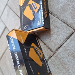 Dvě krabice s produktem BeamZ DMX TRANSCEIVER. Balení jsou černé a oranžové s nápisy „7 UNIVERSES“ a „UNPACK. INSTALL. AMAZE.“. Na krabicích je zobra…